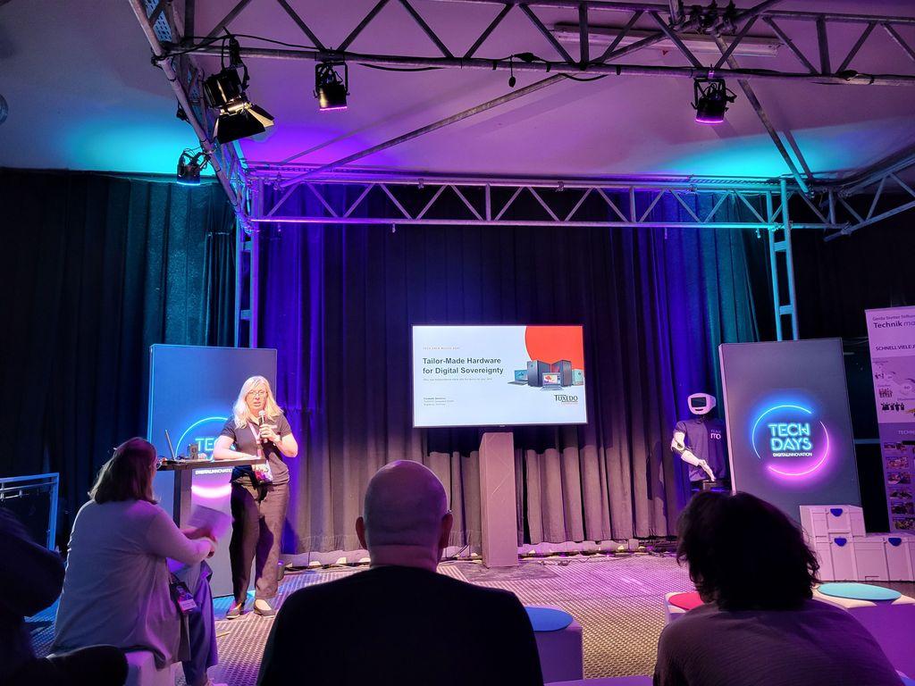 Woman holding a microphone stands on a stage in front of a presentation screen with audience seated in front, stage lit with lighting and "TECH DAYS" illuminated signs.