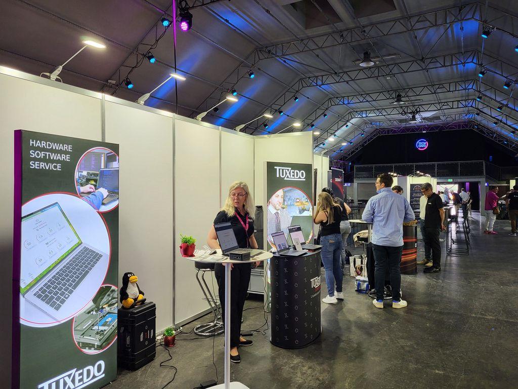 Exhibition booth with two counters where people stand, with more booths and visitors in the background inside a large hall with a vaulted ceiling.