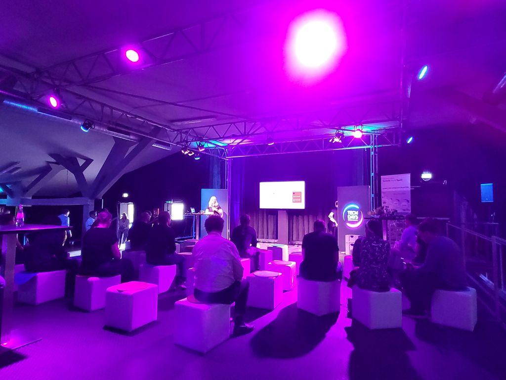 Several people sit on white cube seats in a room lit with purple lighting facing a stage with a screen and a speaker.